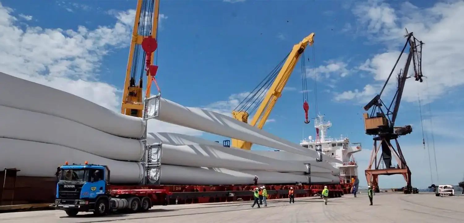 Transporte marítimo y descarga de palas o aspas de aerogenerador (turbina eólica) con grúas amarillas en un camión Scania en el puerto.